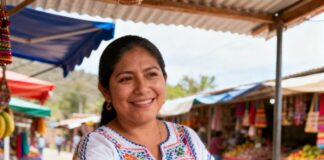 Mujer mexicana adulta en un mercado rural de México, en un puesto pequeño de frutas o artesanías. Ella sostiene un smartphone y muestra una app de pago digital en la pantalla, cobrando a un cliente.